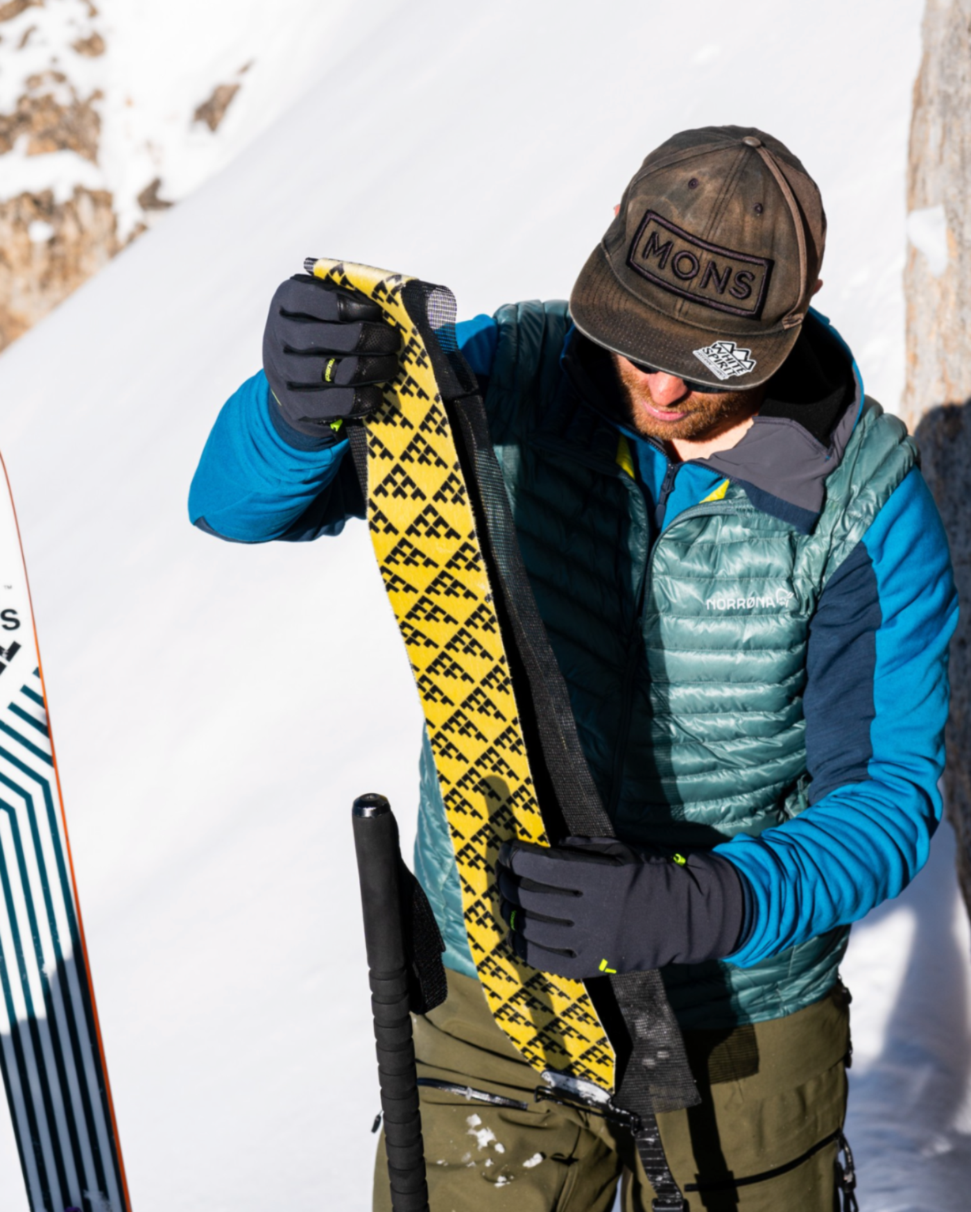 A person in winter gear stands in the snow holding a yellow and black patterned ski binding, with a ski and poles visible nearby.